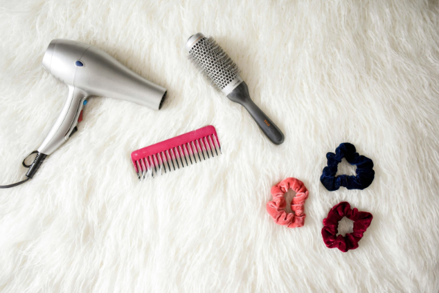 A hair care flat lay featuring a silver blow dryer, a pink comb, a gray round brush, and three scrunchies in navy, blush pink, and burgundy, all arranged on a soft white faux fur background.