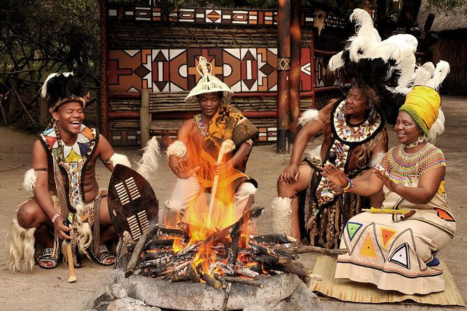 Four people in traditional attire gather around a fire, smiling and drumming. Vibrant patterns and colors accent the cultural setting.