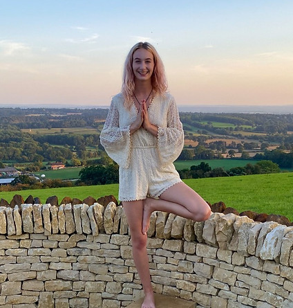 Neurodivergent yoga teacher amy nairne standing in tree pose hands in pray position. Wearing a white playsuit with pink dyed hair.