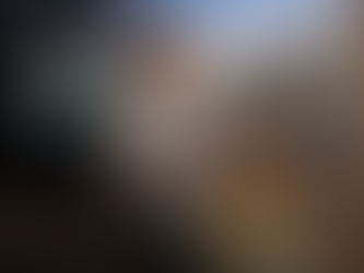 A professional shot of Ariel Flax presenting at the Smart Estate Security Conference. Ariel is standing in the foreground, wearing a white button-down shirt and grey trousers, gesturing with his hands while speaking.  In the blurred background, a large screen displays a presentation slide titled "The Shift: From Reactive to Resilient" alongside an image of a modern estate gatehouse at dusk. The setting has an industrial feel with a visible brick wall to the left.