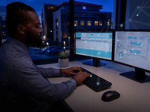 A professional sitting at a desk at night reviewing ATG Digital access control and visitor analytics dashboards across dual computer monitors, with glowing digital network nodes overlaid on the screens.