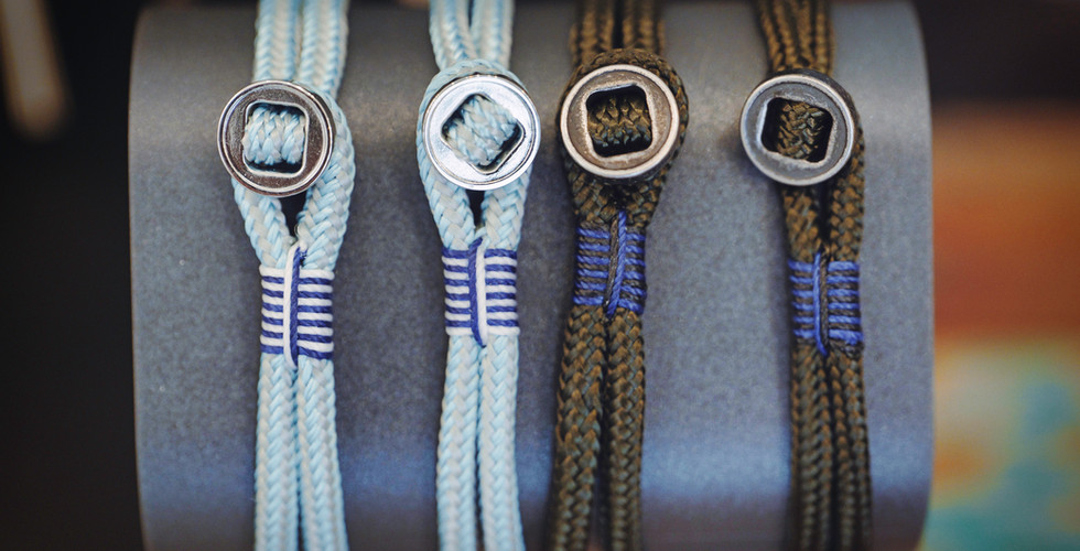"Four Pig and Hen men's rope bracelets in light blue and olive green displayed on a dark grey stand, showcasing stainless steel shackle clasps and detailed colored thread whipping."