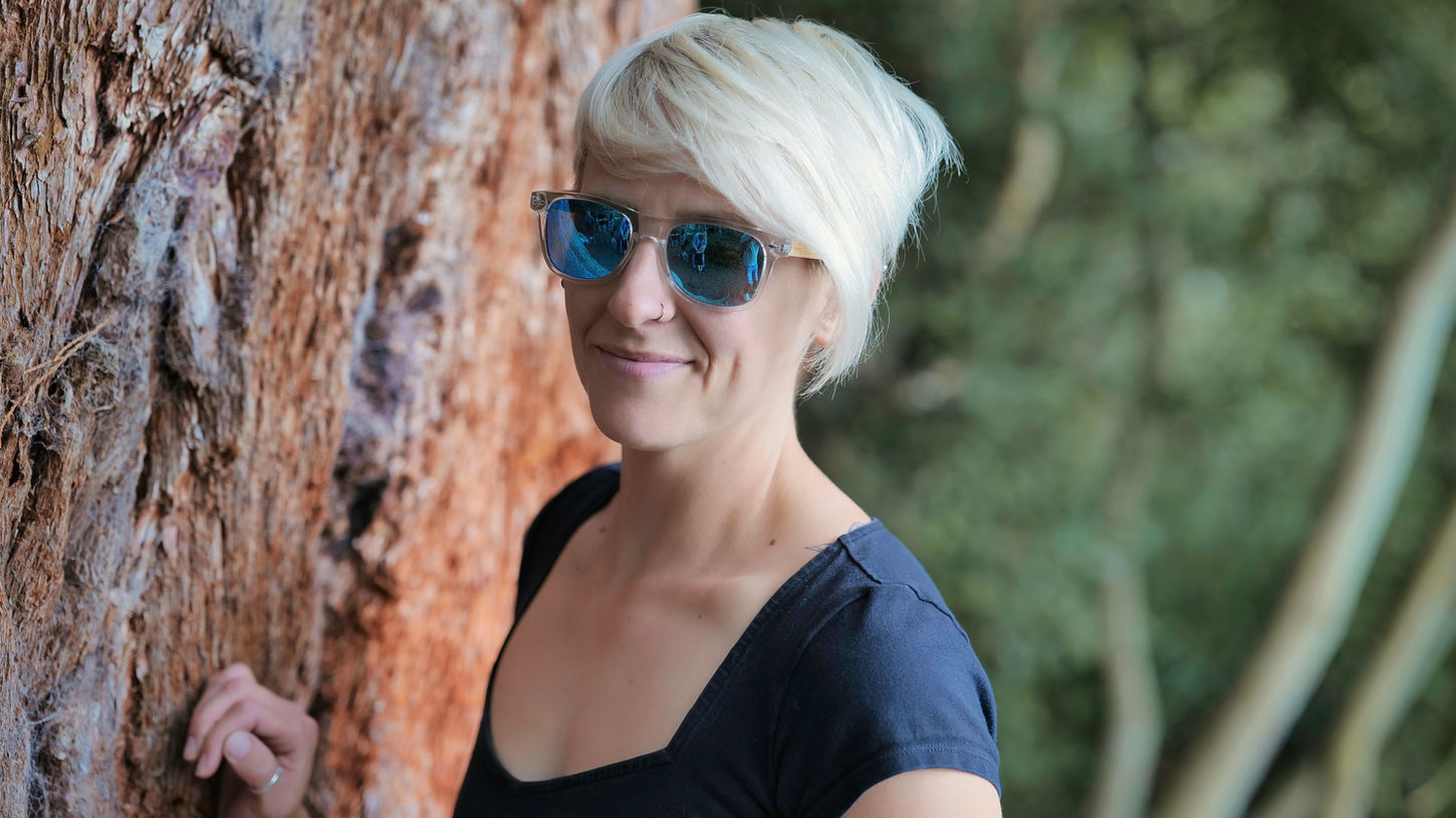Blonde woman in sunglasses posing by a large tree trunk