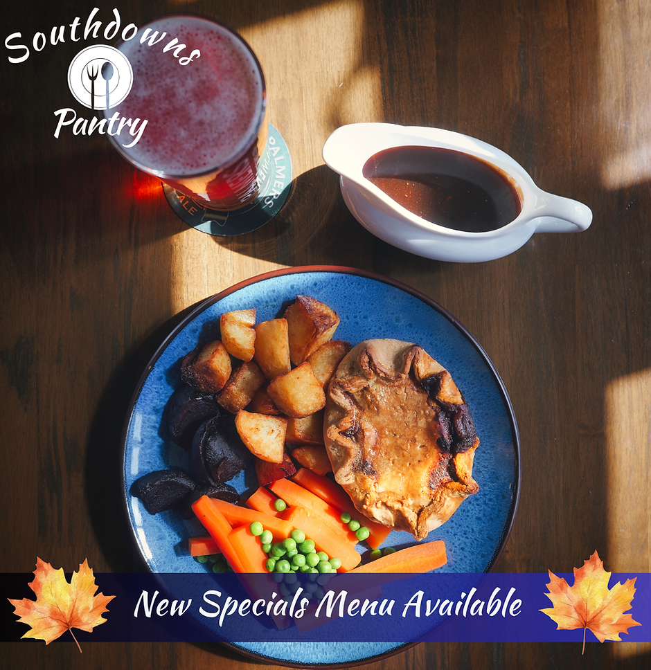 Overhead view of a golden-brown homemade steak pie on a blue ceramic plate, served with roasted potatoes, sliced carrots, peas, beetroot, a boat of gravy, and a pint of Palmers ale on a wooden table.