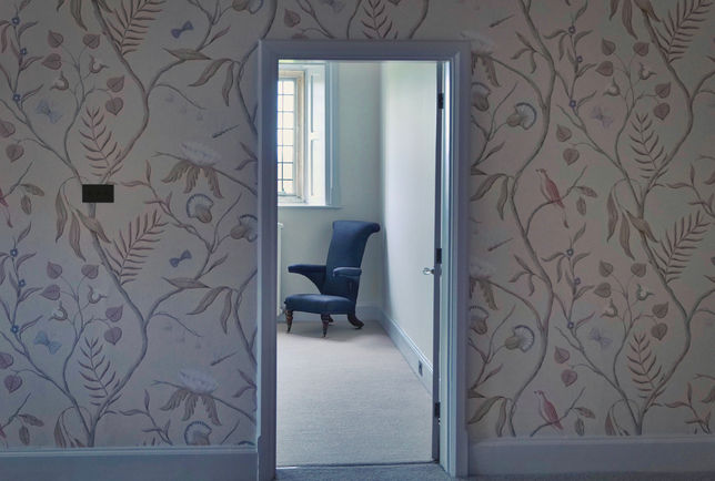Open doorway revealing a room with a blue armchair, a window, and floral wallpaper