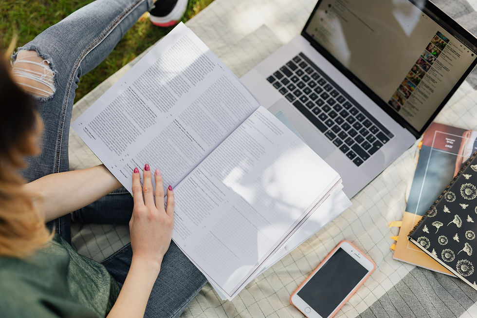 Student reading an open textbook beside a laptop and phone on a picnic blanket outdoors.