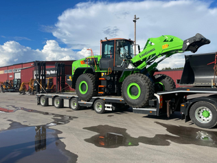 Grön Liugong hjullastare lastad på trailer framför Stig Machines anläggning under blå himmel.