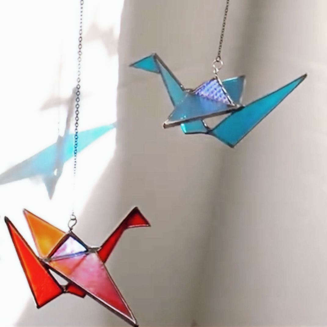 Handcrafted glass origami crane in turquoise Bullseye stained glass, displayed on a windowsill