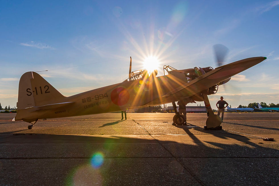 Vintage aircraft on tarmac at sunset, propeller spinning. Sunlight creates lens flare. Two people stand nearby. Tail number S-112 visible.