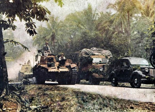 Tank and vehicles move down a tree-lined road in a jungle setting, surrounded by lush greenery. The scene appears calm yet tense.