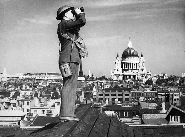 Man in suit and helmet uses binoculars on a rooftop, overlooking cityscape with a large domed building in the background. Black and white image.