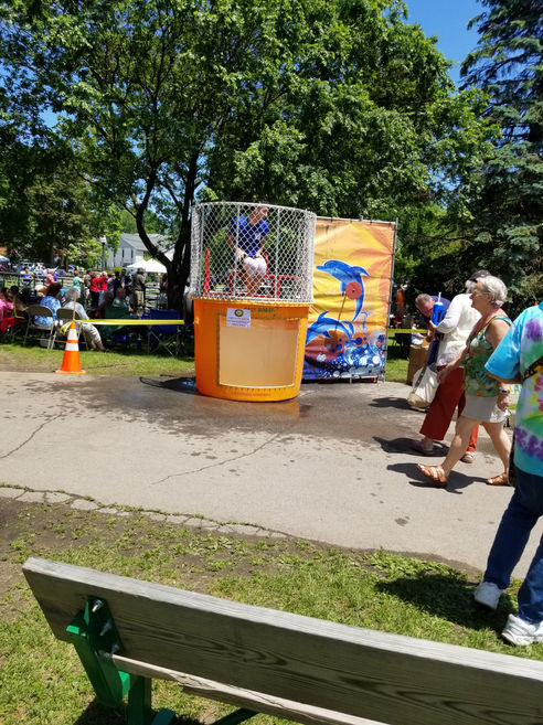 An orange dunk tank with a dolphin backdrop setup on a paved walkway in a public park during a summer member festival.