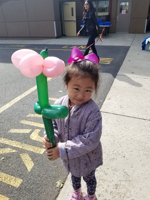 A little girl wearing a bow in her hair holds a large pink and green balloon flower while smiling outdoors.