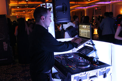 DJ mixing music on laptop and controller at a glowing white booth during prom night with dancing guests in the background.