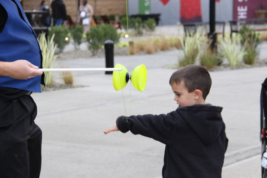 A child extends his arm while a performer demonstrates a diabolo trick with yellow juggling props outdoors.