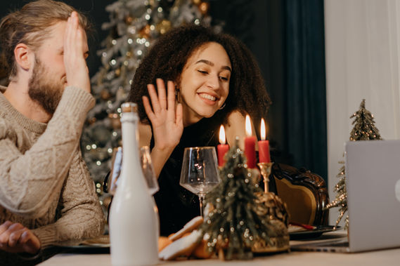 Smiling couple sitting at a holiday-decorated table, waving toward a laptop during an online murder mystery event.