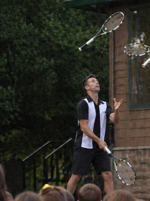 A professional sports juggler actively tossing and catching multiple full-sized tennis rackets mid-air during a live event.