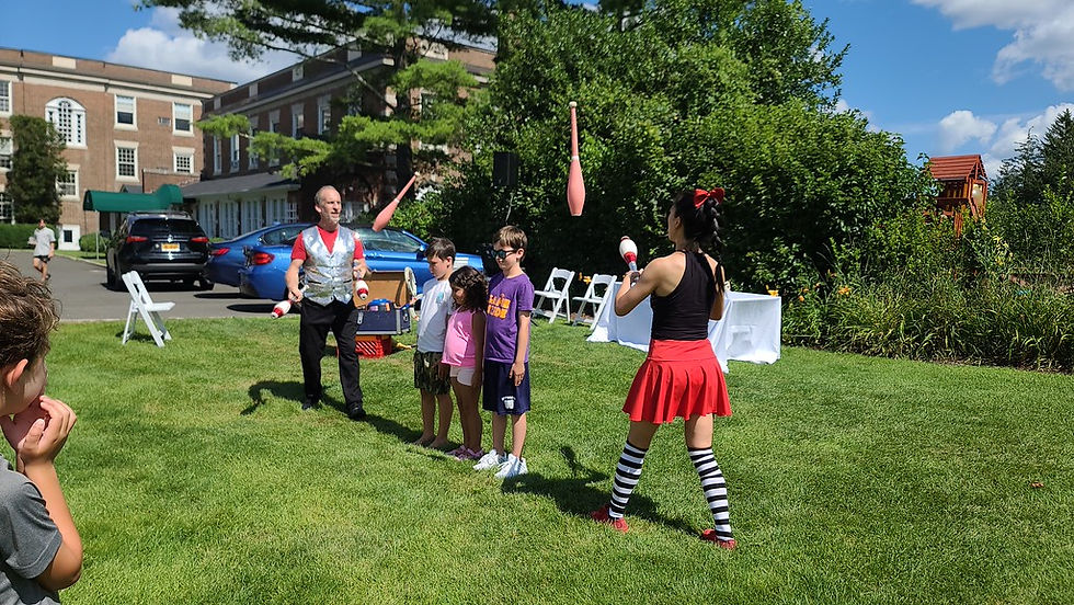Three children participating in an outdoor circus activity with two performers demonstrating juggling clubs.