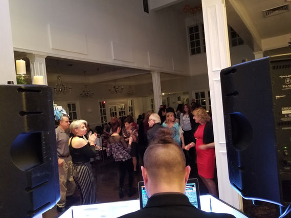 Adults dance and party under colorful lights as a DJ performs on stage at a lively indoor event.