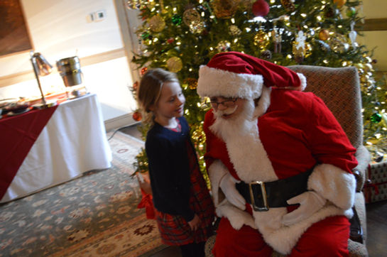 Child talking to Santa Claus while standing in front of a Christmas tree at a holiday event.
