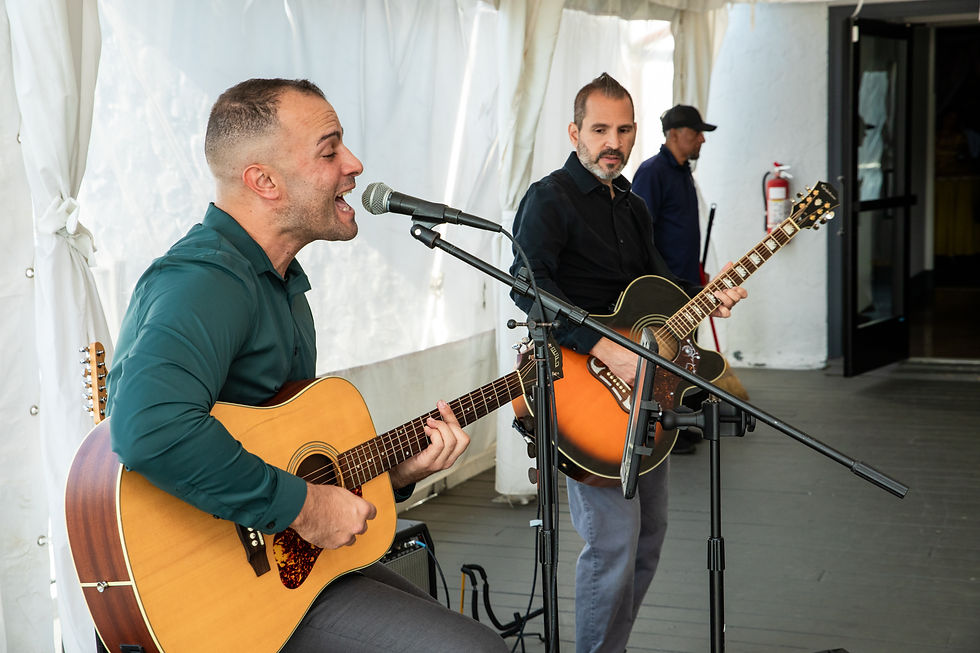 Two acoustic guitarists performing live at an event, with one singing into a microphone while the other plays guitar.