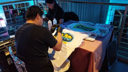 A student getting an airbrush design applied to their apparel at a college event, with an artist using an airbrush tool and vibrant paint