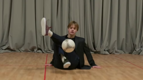 NickFreestyle balancing a soccer ball on his foot while performing a freestyle trick on an outdoor soccer field.
