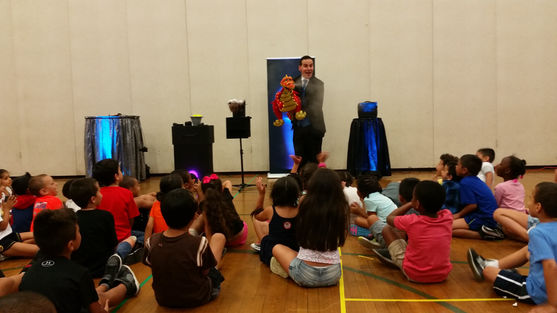 A live puppeteer performing with a smoke-breathing red dragon puppet for children seated in a gymnasium during a Preschool Party.