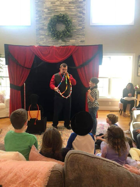 A magician performing an interactive illusion with a child volunteer on stage in front of seated kids at an indoor family reunion.