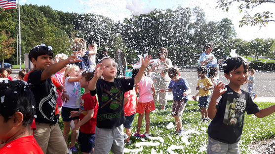 Young kids playing and catching falling artificial snow flurries on a sunny lawn during an outdoor Snow Party event.