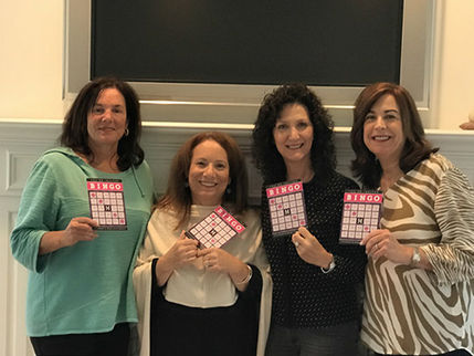 Four smiling women holding pink Glam Bingo cards at a event, standing in front of a large TV display.
