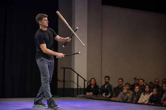 Baseball-themed juggler performing a devil stick trick with a baseball bat on stage.