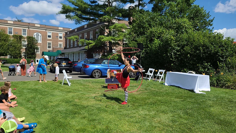 Joyce Kung performing a split on the grass while balancing red hula hoops during an outdoor circus show.