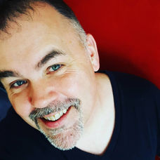 Close-up headshot of Greg R. Baird smiling against a bold red background, wearing a black shirt.