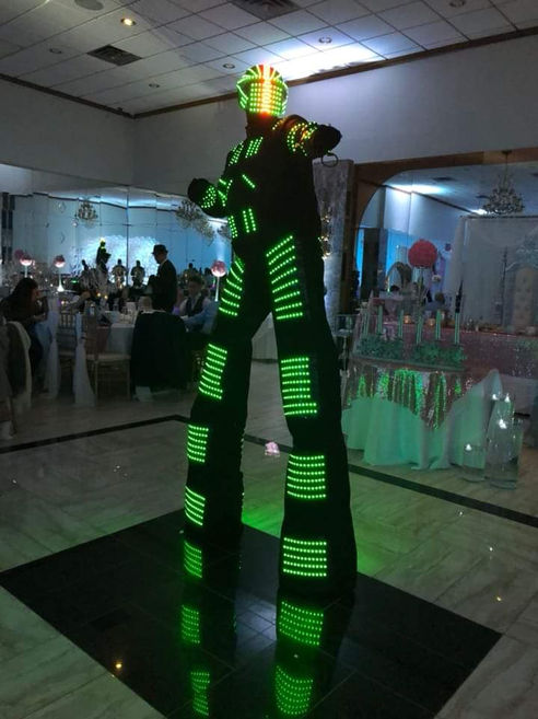 A stilt walker wearing a full-body green LED light suit performing at an indoor family reunion reception.