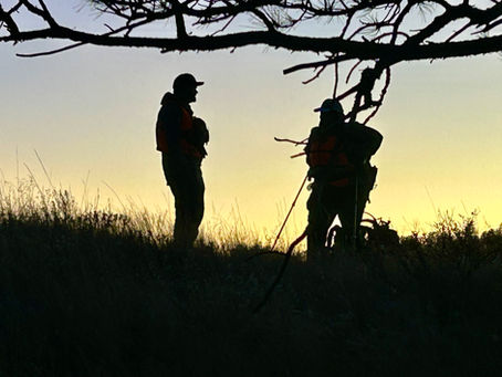 Two hunters pausing on a slope, hands out to feel the shifting mountain wind, attuned to each other and the environment.