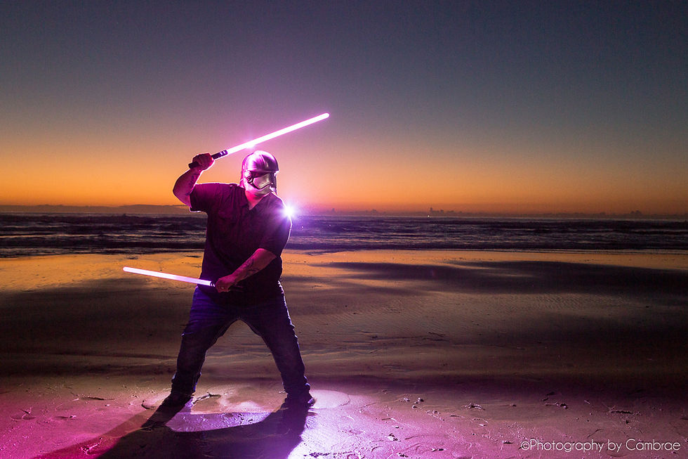 A man with a silver mask on holds pink light sabers on the beach after sunset