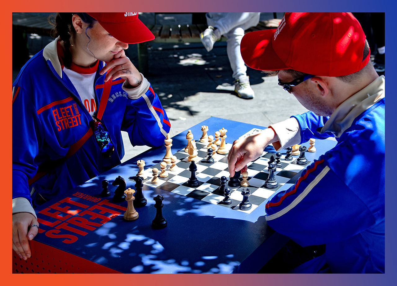 Two individuals dressed in matching blue tracksuits and red caps are engrossed in a game of chess at an outdoor table in Fleet Street Quarter. The table is branded with "Fleet Street" in red lettering.