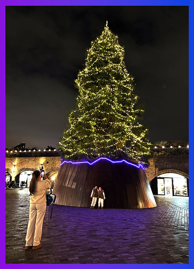A majestic Christmas tree glowing with warm yellow lights against the night sky, with people admiring its beauty and taking photos nearby.
