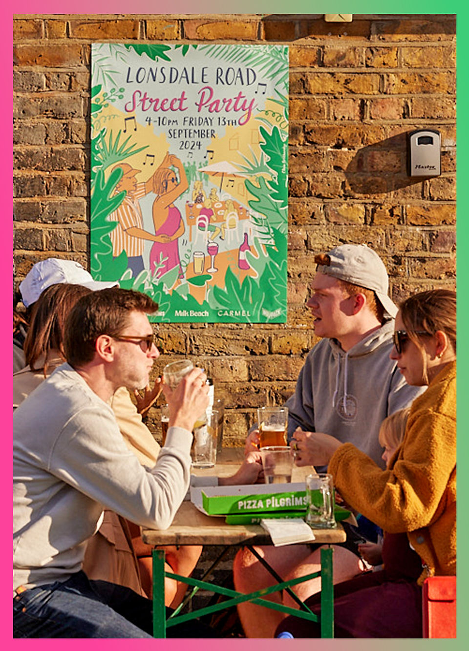 People enjoying pizza and drinks at an outdoor table during the Lonsdale Road Street Party, with a colorful poster promoting the event on a brick wall behind them.