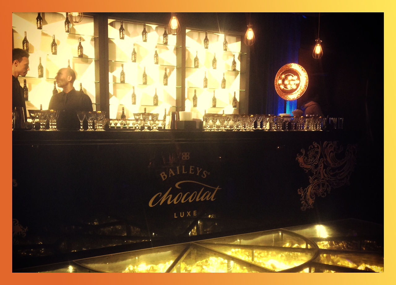 A luxurious Baileys Chocolat Luxe bar setup with shelves showcasing bottles of Baileys, golden-lit decor, and a sleek counter, radiating sophistication