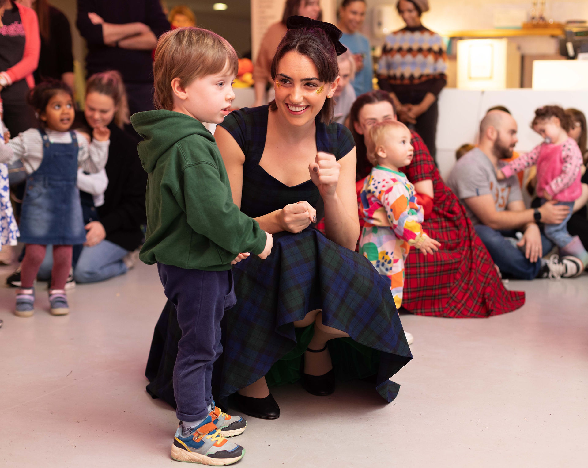 A woman kneels down to engage with a young child in a green hoodie, while other children and parents participate in a lively indoor event.