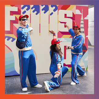 Three individuals dressed in matching blue tracksuits and red caps pose enthusiastically in front of a colourful mural wall created by Kit&Caboodle at Fleet Street Quarter. The mural prominently features the word "FUSION" with vibrant graphics. The group strikes playful poses, adding to the lively and fun atmosphere of the event.