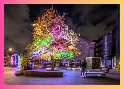 A stunning aerial view of Coal Drops Yard at night, illuminated by thousands of lights. The lights highlight the architectural features of the buildings, with a 45ft Christmas tree standing as a centerpiece.