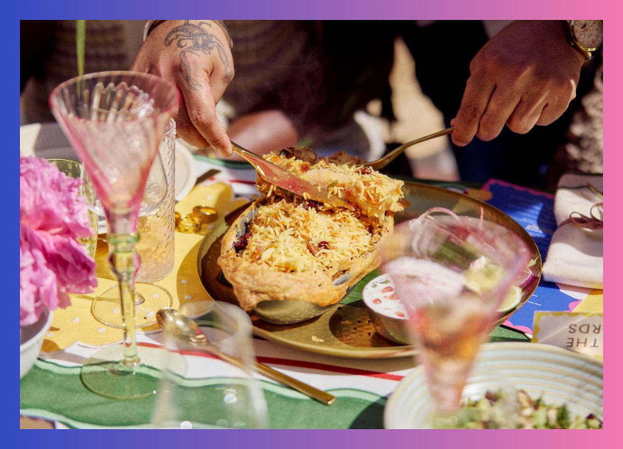 Hands serving fragrant biryani from a pastry crust, surrounded by glassware and colourful table settings.