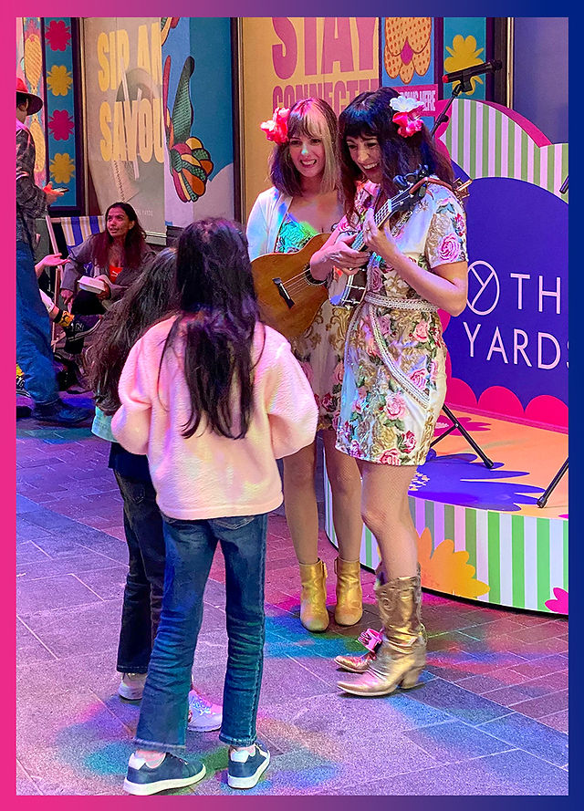 Two female performers with guitars and flower hair clips chat with a group of young children in front of the stage.