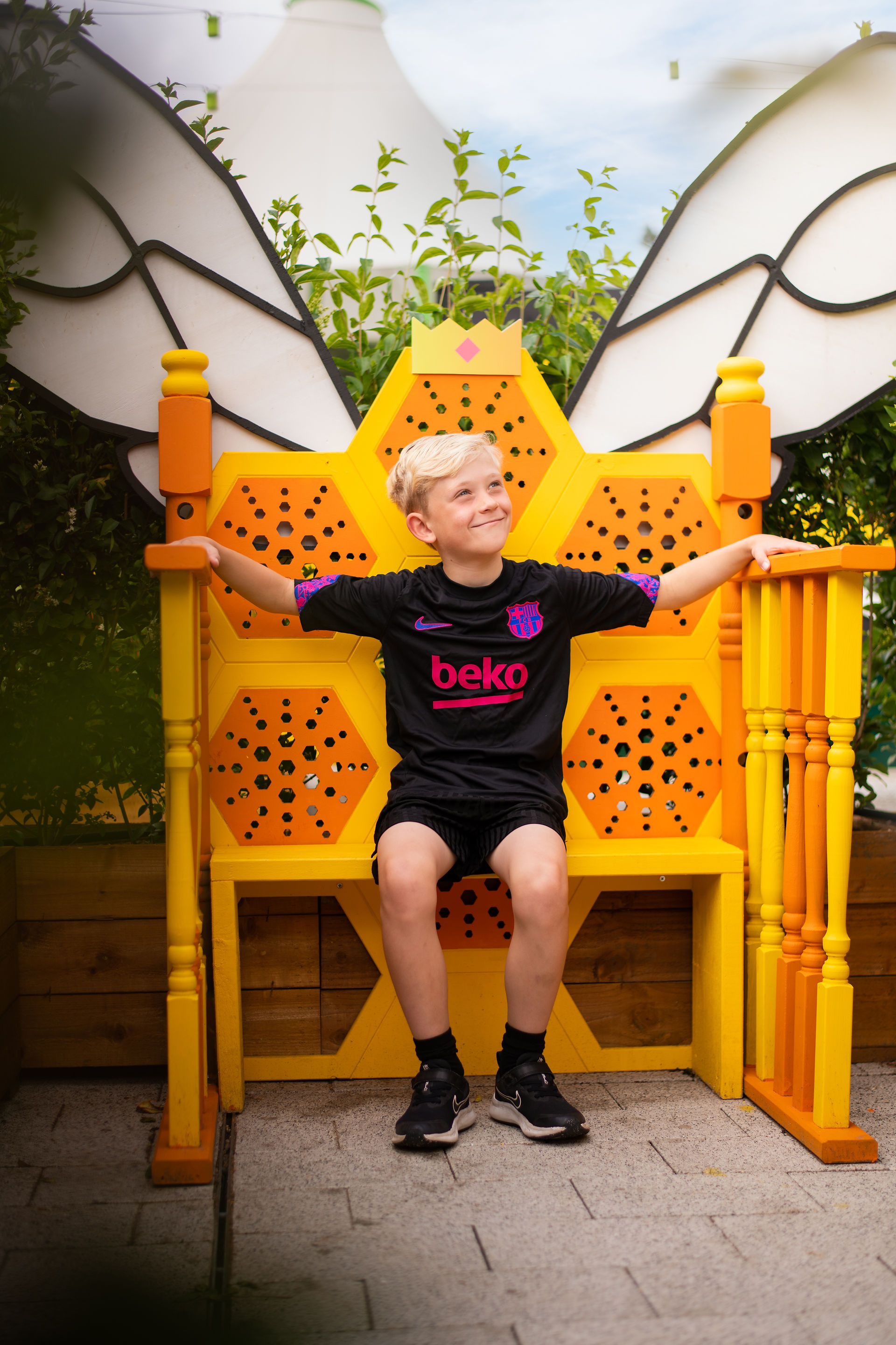 A kid sitting on a throne next to a large illustration of a female bee at the Honey Bee Maze, with visitors exploring the scent-releasing crown feature, designed by Kit and Caboodle, Architects of Happiness.