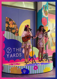 Two female musicians sing and play guitar and banjo on the branded stage, dressed in floral outfits.