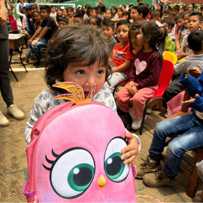Niña sonriendo con mochila rosa, rodeada de niños en evento escolar.