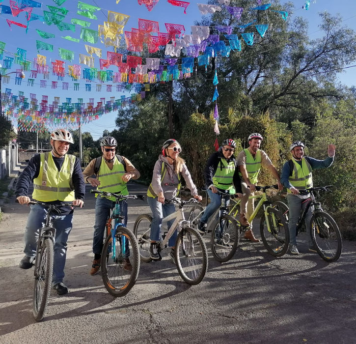 Grupo de ciclistas con chalecos amarillos bajo coloridas decoraciones festivas y árboles.